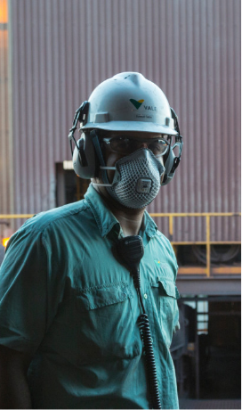 A worker wearing a white safety helmet with the Vale logo, protective glasses, and headphones. He is dressed in a green shirt and has a radio attached to his shirt. In the background, there is an industrial structure with a yellow railing.