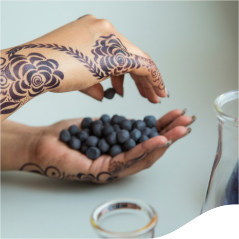 The image shows a person's hands with intricate henna designs holding a handful of blueberries. A glass container is partially visible on the right side of the image.