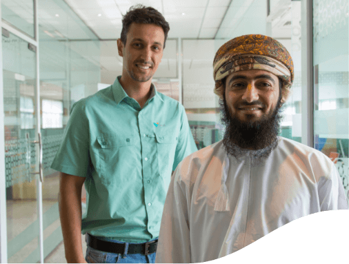 The image shows two individuals standing in what appears to be an office or professional setting with glass walls and doors. The faces are blurred for privacy. One person is wearing a light green short-sleeved shirt and jeans, while the other is dressed in traditional attire with a head covering.