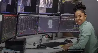 Smiling woman seated in front of multiple monitors displaying satellite and city images
