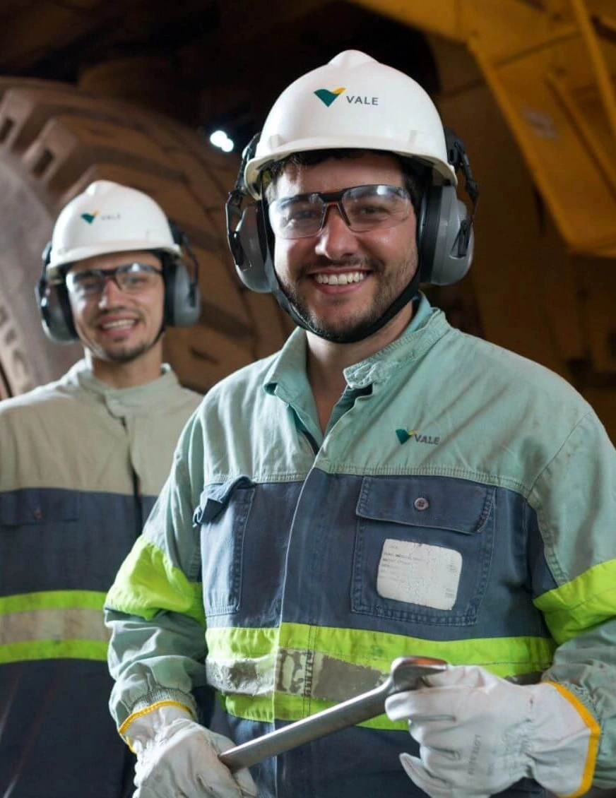 Two Vale employees with white helmets and ear protection smiling at the mining facility