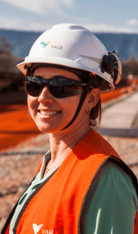 Photo of woman smiling taken from the chest up an operational area. She is wearing a light green shirt, an orange vest, goggles and a white helmet with Vale logo.