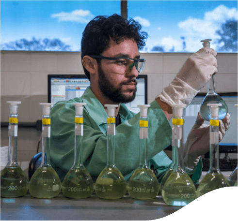 Homem dentro de um laboratório com vários recipientes de vidro a sua frente. Ele usa óculos de proteção e luvas. Está segurando um dos recipientes de vidro nas mãos e ao fundo há computadores.