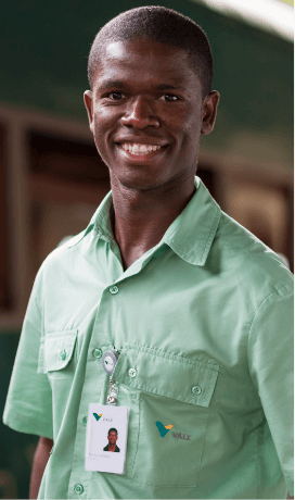 Homem negro sorrindo em uma área operacional. Ele usa camisa verde clara e um crachá.