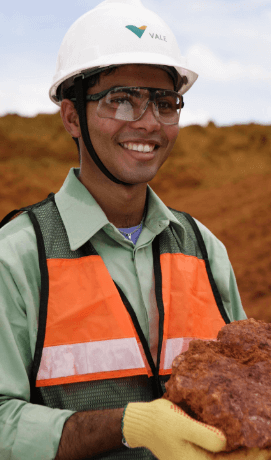 Homem sorrindo em um espaço terroso de operações. Ele segura uma rocha nas mãos e usa camisa verde clara, colete com detalhes em laranja, luvas amarelas, óculos de proteção e um capacete branco com logotipo da Vale.
