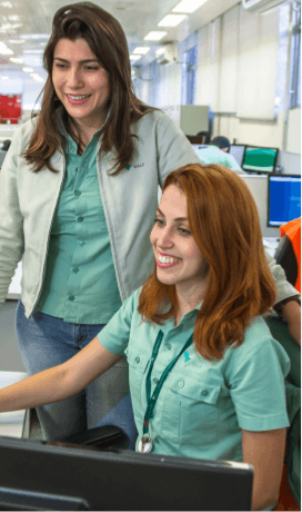 Duas mulheres lado a lado – uma sentada e outra em pé – em um espaço de escritório. As duas estão sorrindo, usam os cabelos soltos e camisa verde clara.