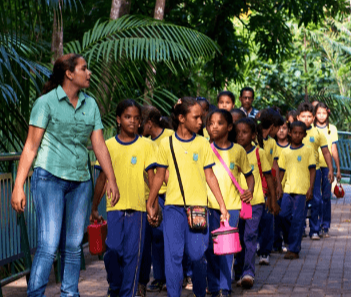 Uma empregada Vale acompanha um grande grupo de crianças, todas vestidas com roupas escolares, por um caminho arborizado.