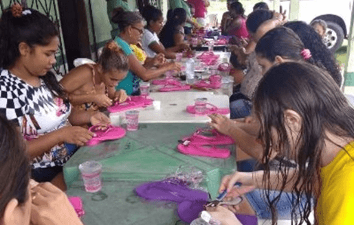 Em uma mesa, diversas mulheres produzem artesanato em chinelos.