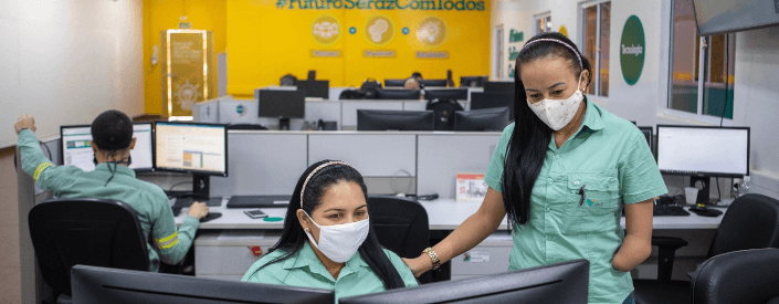Two female Vale employees chat in an office. One is seated and the other is standing. Both are wearing masks.