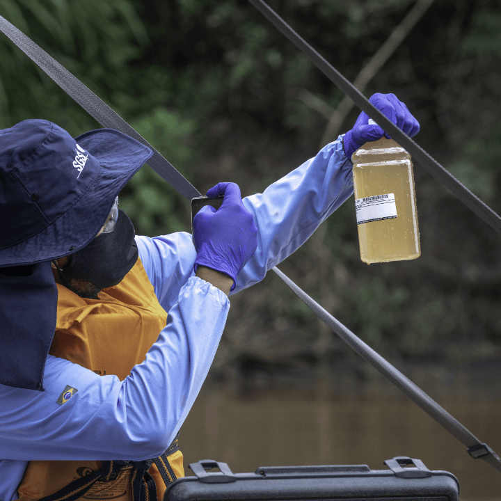 A person, wearing a hat, a mask, a vest and gloves, is holding a glass jar with water taken from a river in one hand, and with the other hand, he is taking a photo of the vessel.