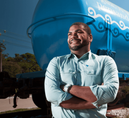 A black man smiling and looking forward. He is wearing a light green shirt with Vale logo. and behind him, there is a blue and white metal structure.