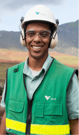 A man is looking at the camera and smiling. He is wearing a green vest with yellow Vale details, a helmet, and goggles.