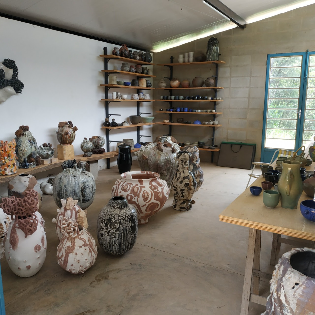 Imagem do ateliê de Benedikt Wiertz. O local possui diversos grandes vasos de cerâmica no chão e ao fundo há prateleiras com objetos menores.