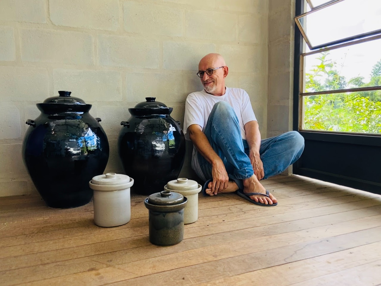 Um homem está sentado no chão ao lado de dois grandes vasos pretos e três vasos menores.