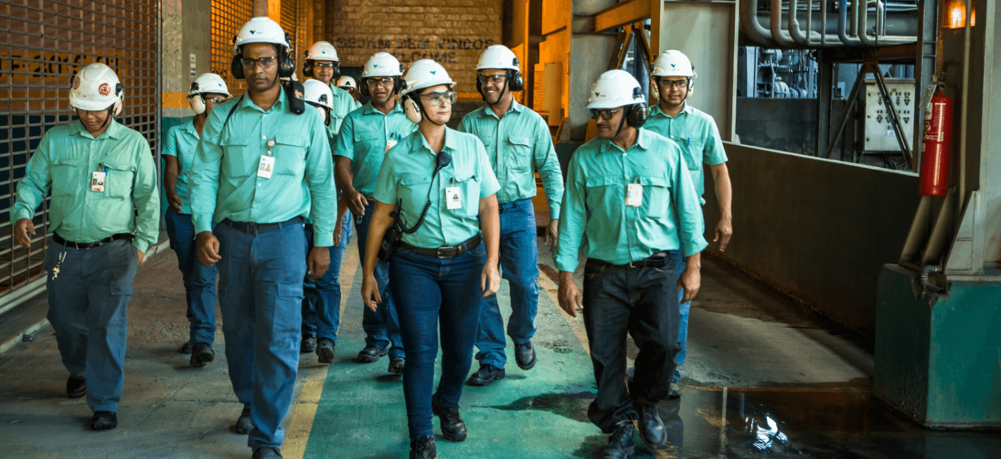 Vários empregados da Vale caminhando lado a lado em um espaço operacional. Todos usam uniformes verdes, crachá, óculos de proteção, protetores de ouvido e capacete branco com logotipo da empresa.