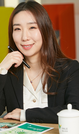 A women smiling. She wears executive clothes and hold a pencil next to her face