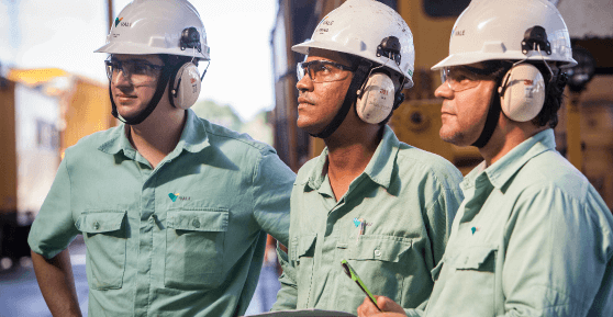 Photo of three men observing a point and a large iron machine in the background. The three are wearing light green button-down shirts with Vale logo, ear protection, helmets and goggles. One of them is holding a pen and paper in his hand.