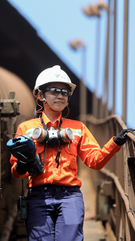 A Vale employee in the operations area. she is wearing an uniform, goggles, gloves and a protective hat, while holding a piece of equipment in her hand.