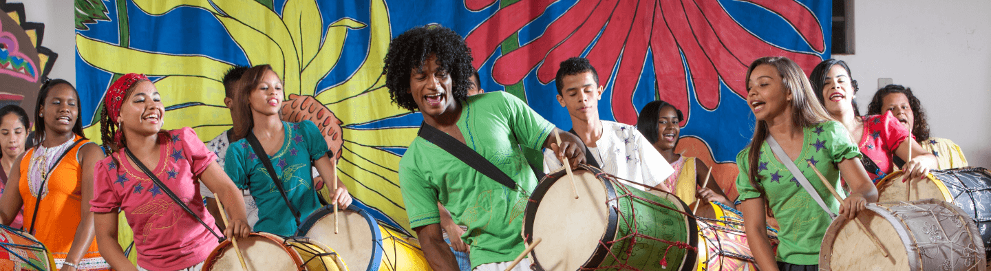 Young people play drums and smile, as if they were singing. At the back there is a fabric printed with the colors blue, red, yellow, and green.