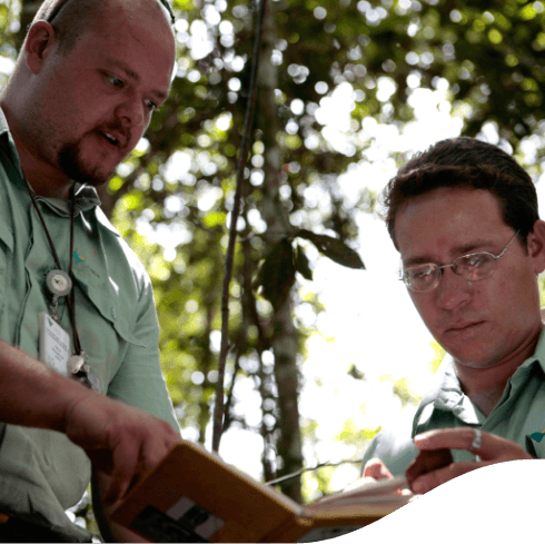 Two men stand side by side in a tree-filled space. They both wear uniforms in a light shade of green and are evaluating what is written in a notebook.