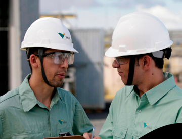 Two men talking in an operation area. One of them is holding a clipboard. Both are wearing light green Vale button-up shirts, goggles and helmets.