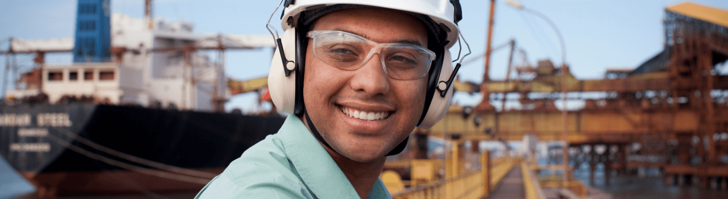 Man smiling in an operations space with a metal structure and a ship in the background. He is wearing a light green shirt, goggles, ear muffs, and a white helmet.