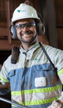Vale employee smiling in an operational space. He is wearing light green uniform, radio communicator fixed to the shoulder, goggles, ear muffs and a white helmet with Vale logo.