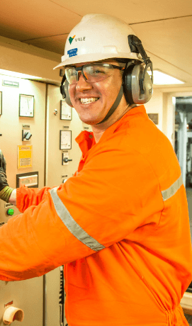 A man smiling in an operational area. He is wearing orange uniform, goggles, ear muffs and a white helmet with Vale logo.