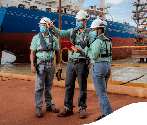 Vale employees standing side by side in a company port. All wear uniforms with gray pants and green shirts, face masks, goggles and  white helmets.