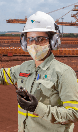 Vale employee in an operational area holding a radio communicator in one hand. he is wearing green uniforms, a face mask, goggles, ear muffs and white helmets with Vale logo.