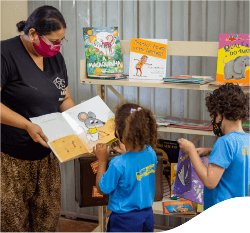 A woman wearing a black T-shirt and face shield shows a children's book to a girl in front of her. The girl is dressed in blue and her hair is tied up. Next to her, there is a boy also dressed in blue. Behind them, there is a shelf with several children's books.