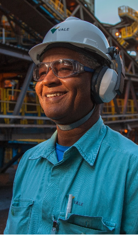 Photo of a man smiling, and in the background, an operation area with several iron structures. He is wearing green Vale uniform, ear protection, helmet and goggles.