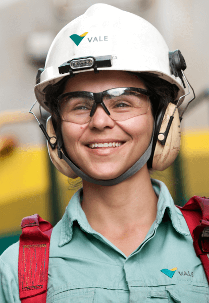A women smiling in an operational area. She is wearing light green shirt with Vale logo, red safety equipment fixed to the shoulders, goggles, ear muffs and a white helmet with Vale logo.