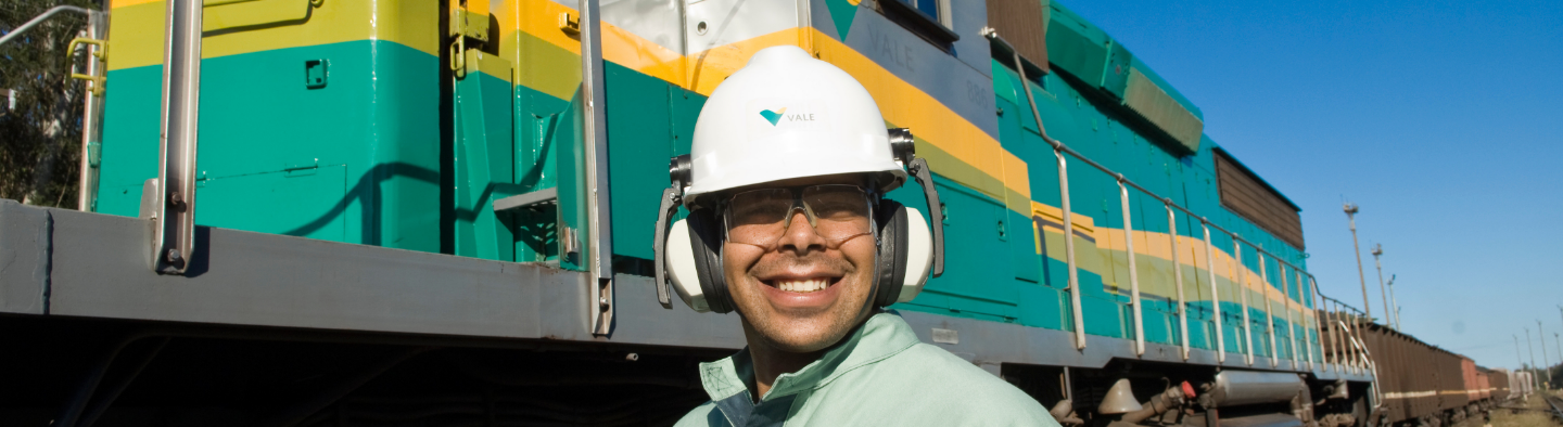 Empregado Vale sorrindo. Ele usa capacete e óculos de proteção. Ao fundo há um trem Vale, nas cores verde e amarela.