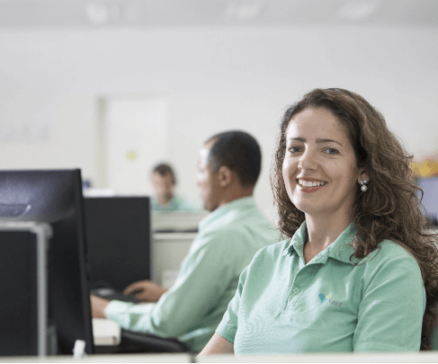 Photo of a woman smiling in an office in front of a monitor. She has wavy hair, wears pearl earrings and a light green polo shirt with Vale logo.