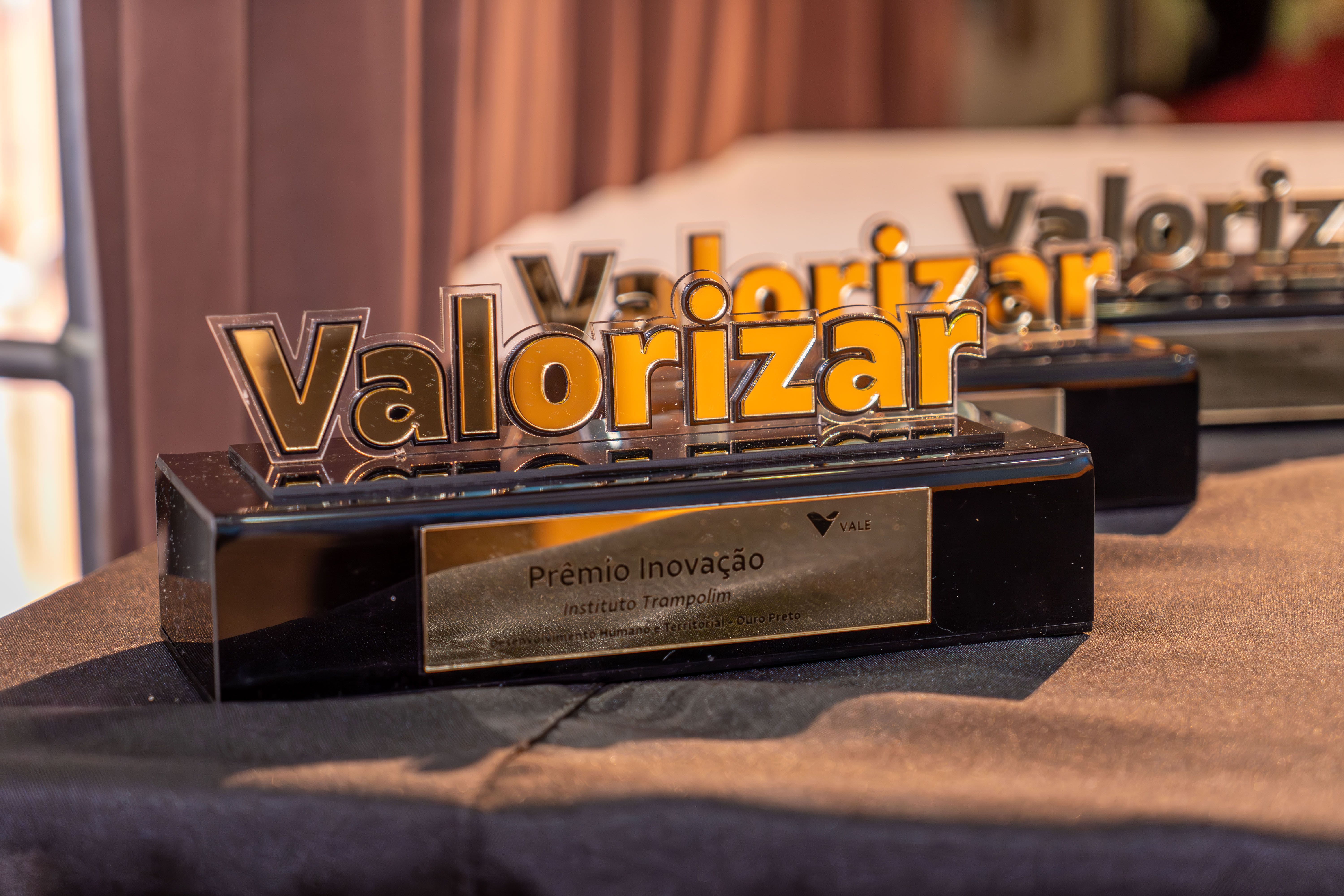 Imagem do prêmio. Em uma estrutura preta há uma placa escrito “Prêmio Inovação” e em cima a palavra Valorizar, em relevo.