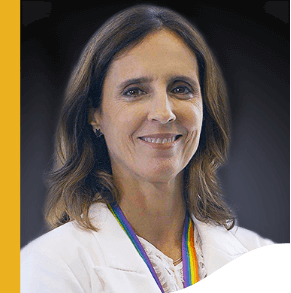 Photo of Marina Quental. She has shoulder-length brown hair, is smiling, and is wearing a white blazer and a lanyard that ties her badge with the colors of the LGBTQIA+ flag.