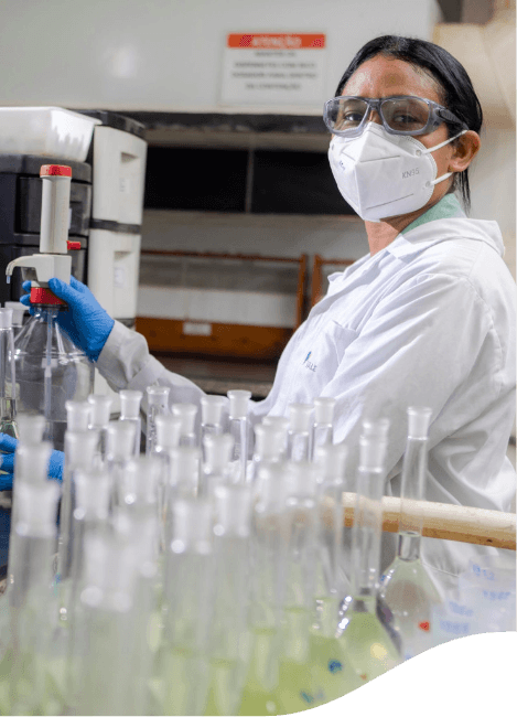 In the foreground are several test tubes. In the background, a researcher looks at the photo. She wears a lab coat, gloves, goggles, and a protective mask.