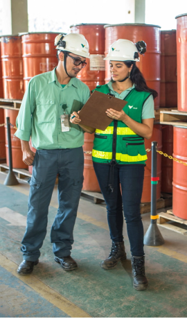 Photo of a man and a woman talking, and in the background, several red barrels. The woman is holding a clipboard and the man is pointing at it. The woman is wearing jeans, boots, a green Vale shirt, a green vest with yellow signs and a helmet. The man is wearing boots, gray pants, a green Vale shirt, glasses and a helmet.