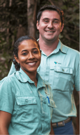 Foto tirada da cintura para cima de dois funcionários da Vale – um homem e uma mulher – sorrindo em uma área arborizada. Os dois usam camisas verdes em um tom claro.