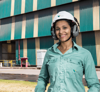 A women smiling in an operational area. She is wearing a light green shirt, ear muffs and a white helmet with Vale logo