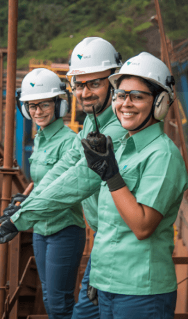 Three employees – two women and one man – stand side by side on a metal structure inside an operation. The three are smiling and wearing light green shirts, goggles, ear muffs and white helmets with Vale logo.