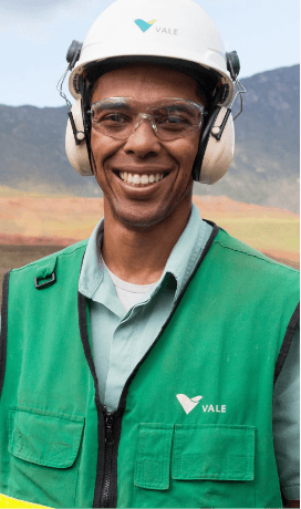 A man smiling in a operational space. He wears a light green shirt and a darker green vest with white Vale logo, goggles, ear muffs and a white helmet with Vale logo.