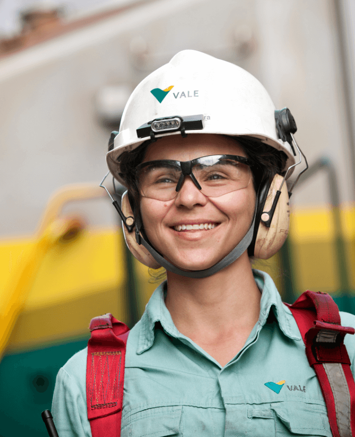 A women smiling in an operational area. She is wearing light green shirt with Vale logo, red safety equipment fixed to the shoulders, goggles, ear muffs and a white helmet with Vale logo.