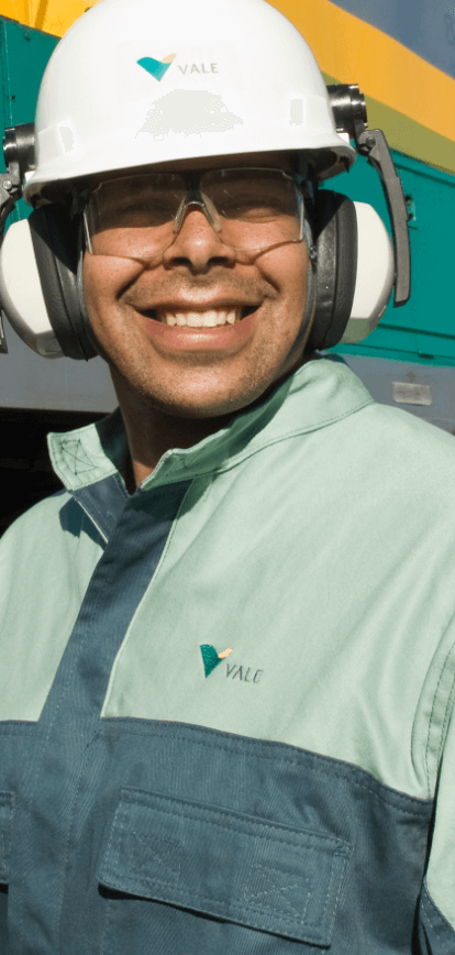 A man smiles for the photo. He is wearing a light green uniform, ear protectors, goggles, and a white helmet with the Vale logo. Behind him is a green and yellow locomotive.