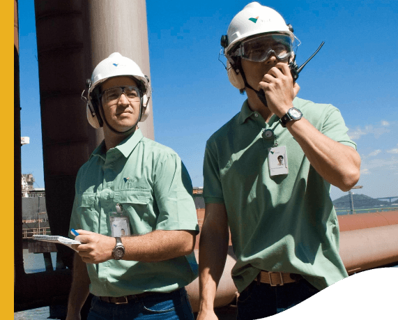 Photo of two men in a high part of an operation observing a point. One of the men is holding and talking on a radio with one hand, he is wearing a light green polo shirt with Vale logo, a badge, a watch, ear protection, a helmet and goggles. The other is holding a notebook and a pen and is wearing a light green button-down shirt with Vale logo, a badge, a watch, ear protection, a helmet and goggles.