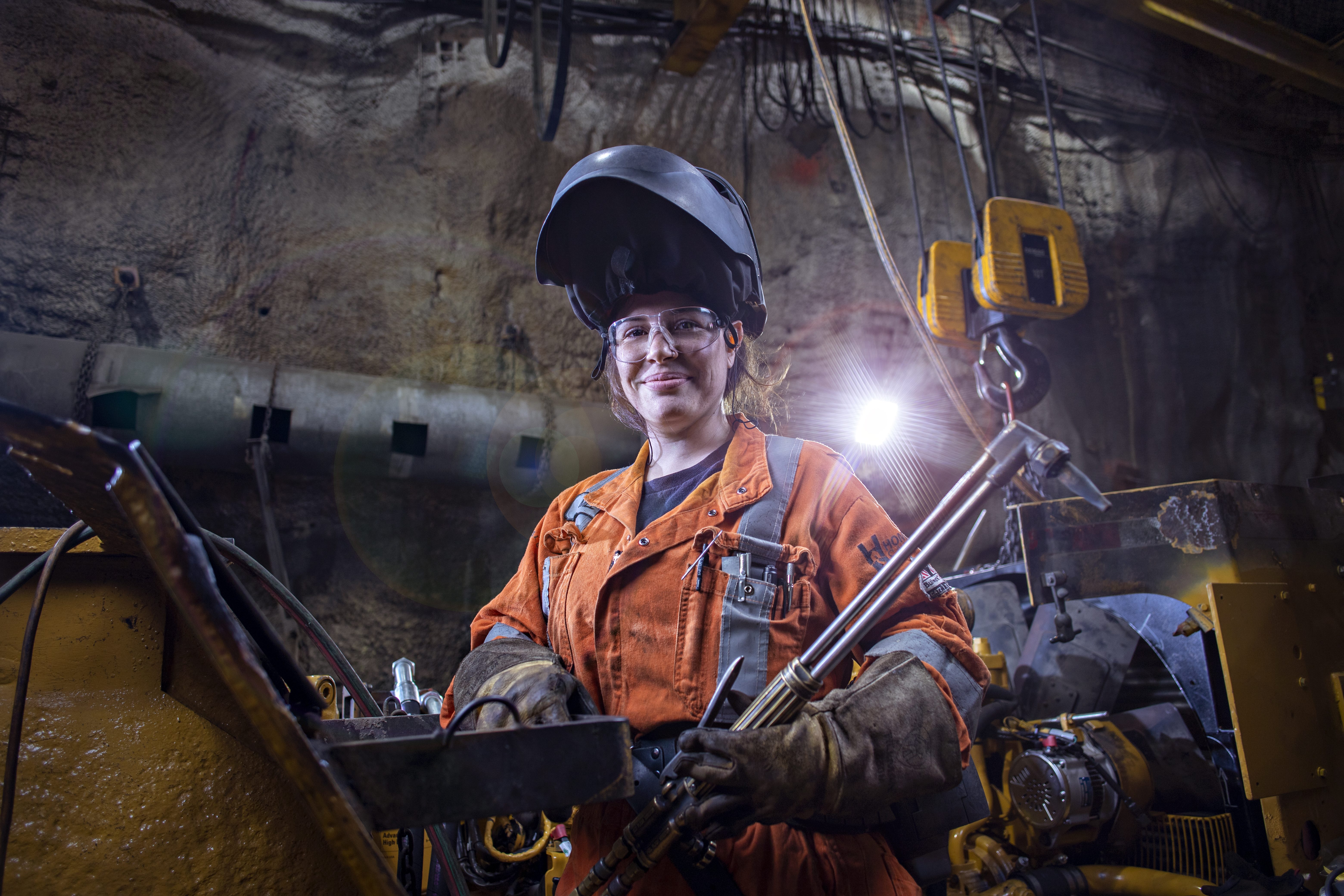 Vale employee with a piece of iron equipment in her hands. She is in an underground space, wearing protective equipment and smiling for the photo.