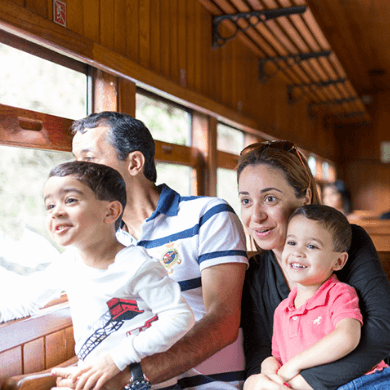 Uma família, composta por um homem, uma mulher e dois meninos pequenos, cada um no colo de um adulto. Todos olham pela janela de um trem.