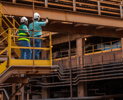 Two Vale employees stand under an iron structure and one of them points upwards. Both are wearing protective helmets.