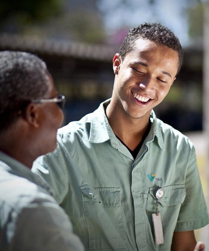 Photo of a black man facing forward smiling and chatting with another black man appearing in profile. The two are wearing the Vale uniform, green button-up shirts